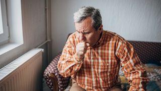 photo of an older man sitting on a couch indoors and coughing into a closed fist