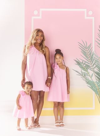 A photo of Serena Williams posing with her daughters in pink one-shoulder sun dresses.