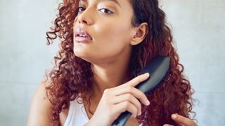 A woman with red, curly hair is pictured brushing the ends of her hair with a wide hairbrush