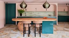 pink kitchen with teal kitchen island with round stone orb, patterned bar stools, crazy paving floors, a mint green marble backsplash and brass pendant lights