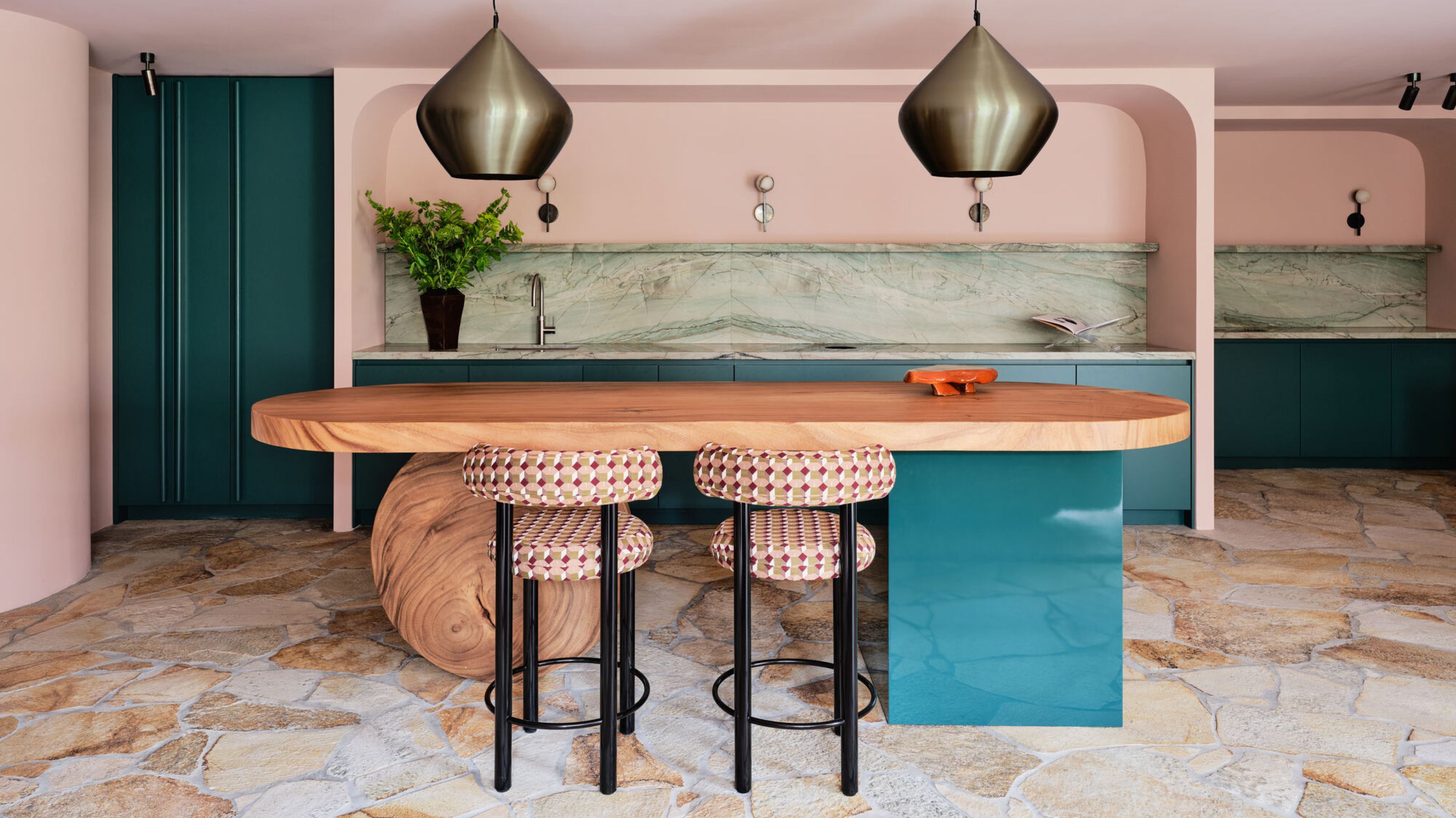 pink kitchen with teal kitchen island with round stone orb, patterned bar stools, crazy paving floors, a mint green marble backsplash and brass pendant lights