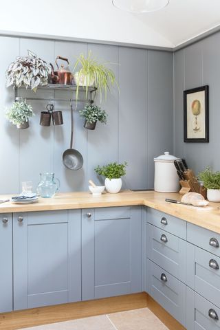 kitchen corner with pale gray blue cabinets wooden worktops and painted paneled walls pale stone floors and plants hanging from wall rack