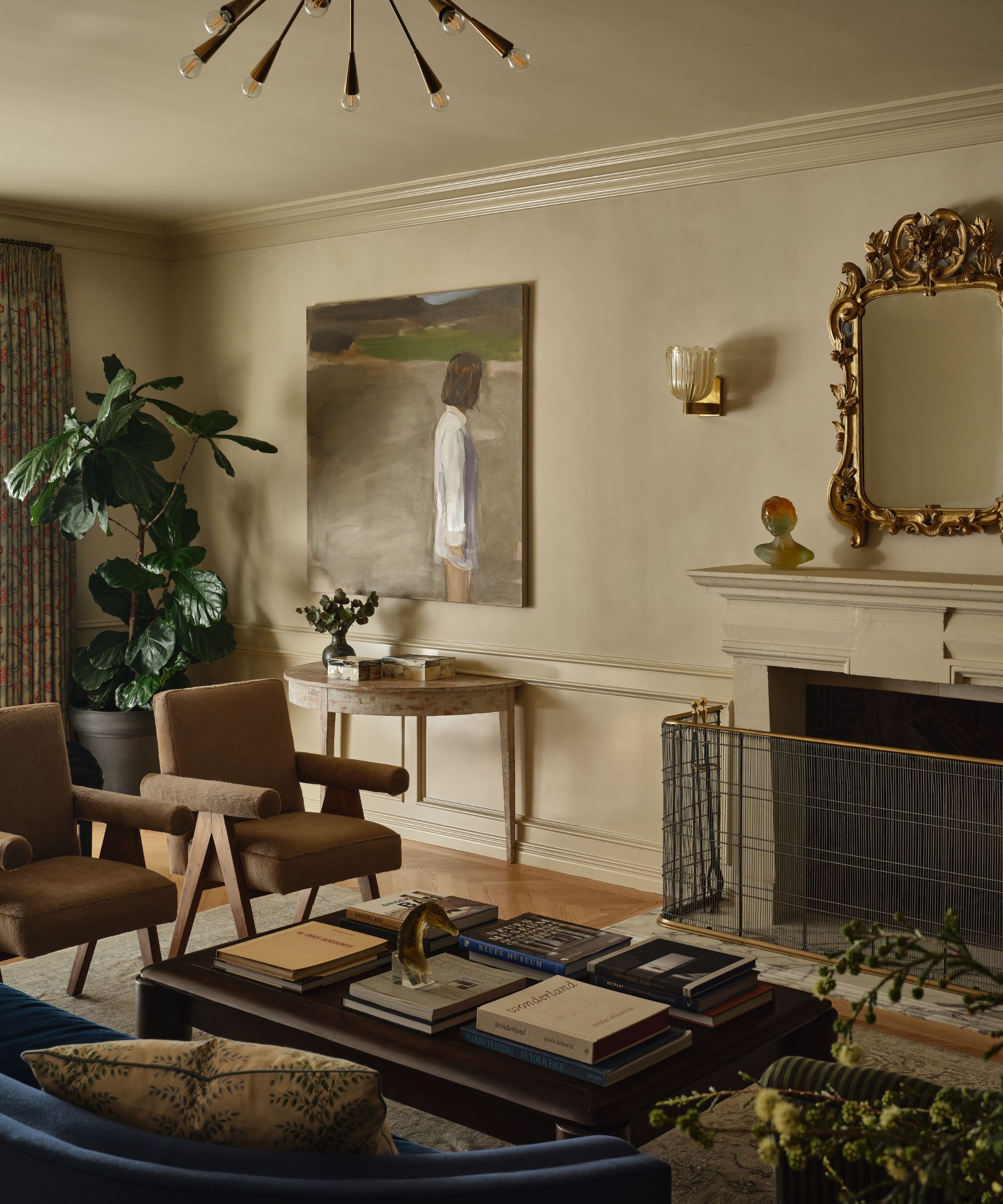 Living room with a classic fireplace, two brown wood-framed armchairs, a dark coffee table covered in books, and a large contemporary painting of a figure
