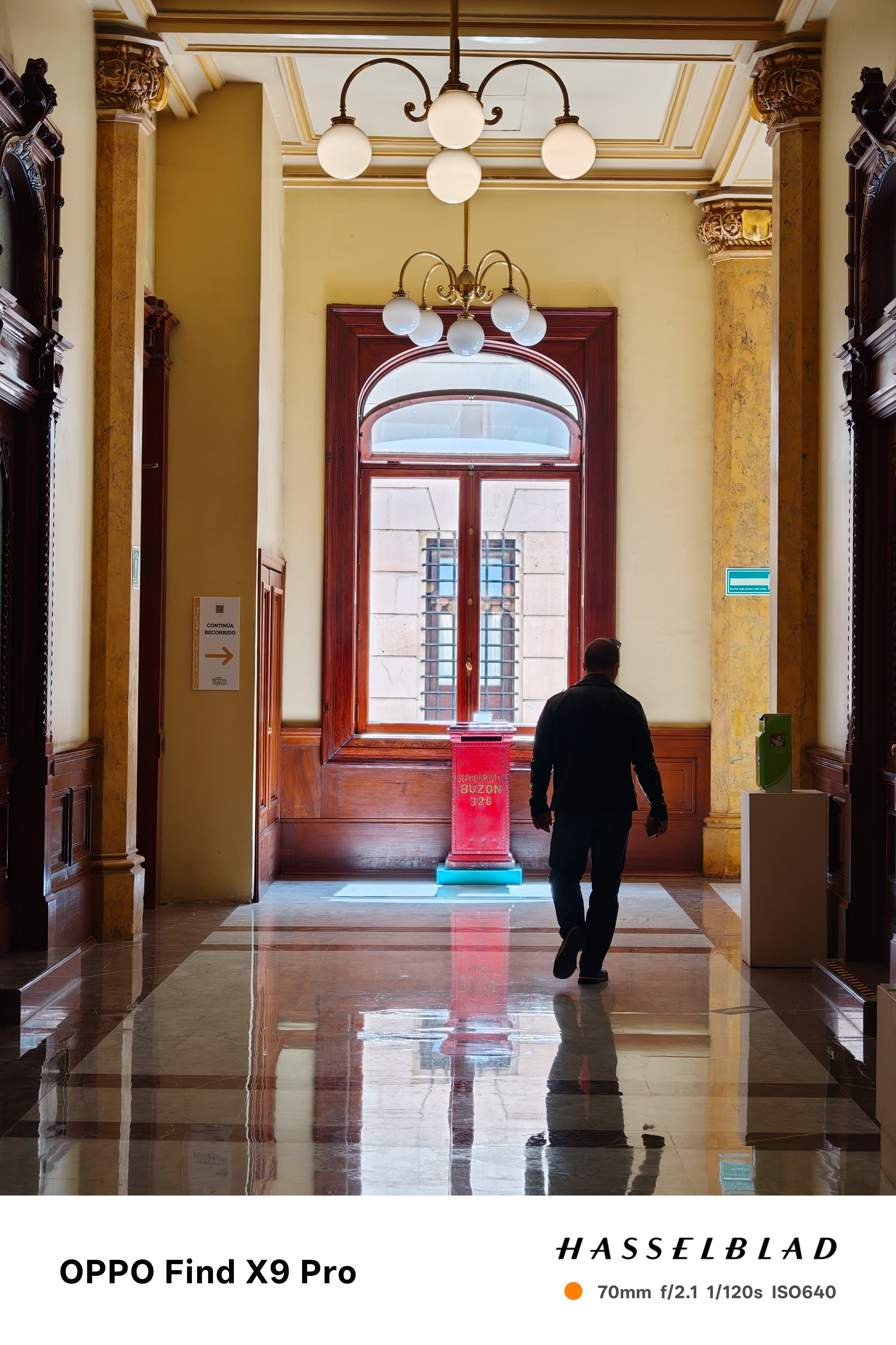 A solid person walks down a corridor in the Palacio postal toward a postbox lit by the sun through a large window
