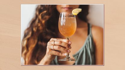 Image of woman holding glass of kombucha with lemon wedge in a white frame against a beige background