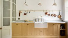 A bright white kitchen with ochre colored cabinets, marble countertops, a gold rail with copper pans and accessories hanging up, three white pendant lights, and a white glass paned door to the left