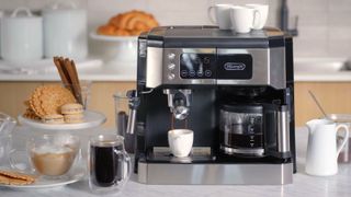 The De'Longhi coffee maker on a kitchen surface surrounded by pastries, biscuits, and a cup of coffee