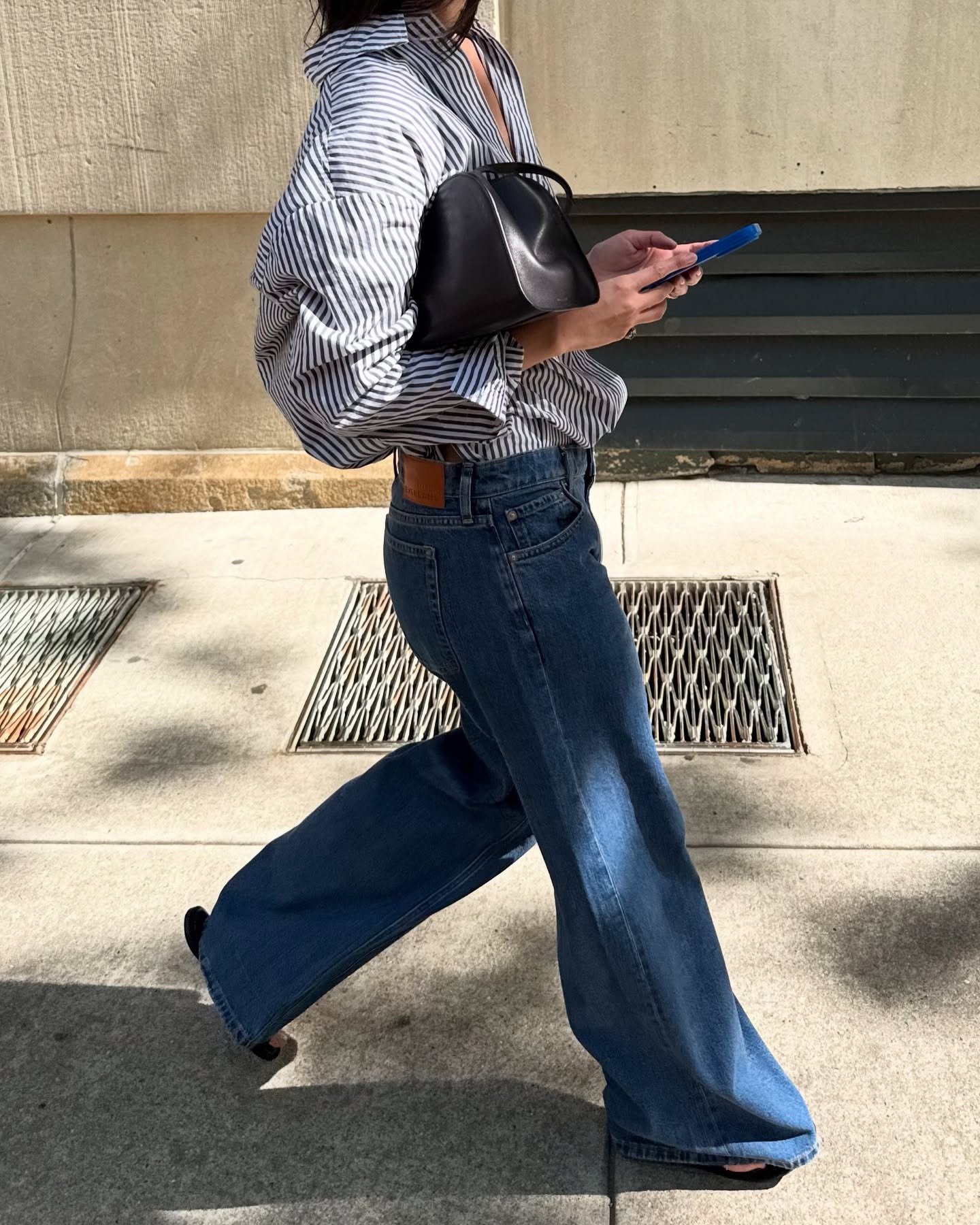 NYC style influencer Mimi Nguyen walking while on her phone wearing an oversize striped button-down shirt, simple black bag, and oversized relaxed jeans