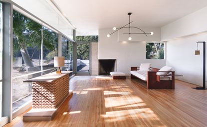 Interior view of Fitzpatrick-Leland House featuring white walls, wood flooring, large windows, a black chandelier, a sideboard with a lamp on top and wooden seating with white padding and cushions