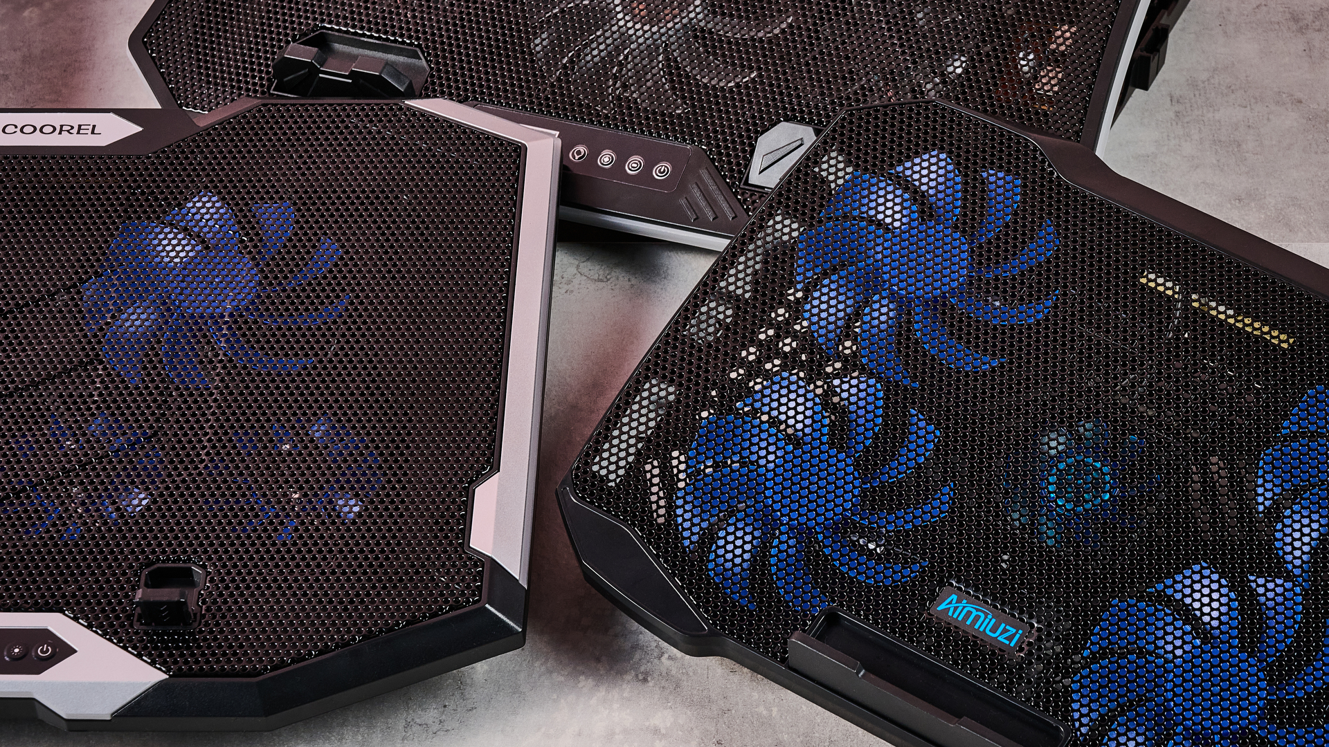 A close up of the TopMate C12, Aimiuzi F2 Plus and ICE COOREL RGB Laptop Cooling Pad on a gray marbled desktop in front of a pink background.