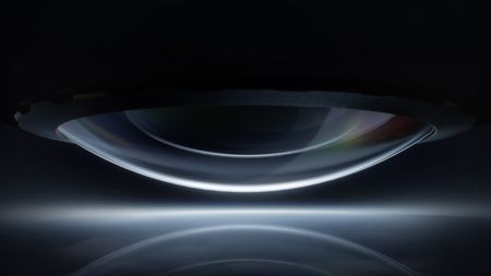 A giant, bulbous camera lens facing down, reflected in light, with low key lighting and a black background