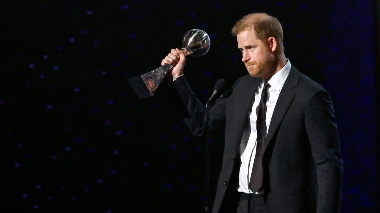 Prince Harry holding up the Pat Tillman Award for Service on stage wearing a black suit 