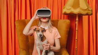 A woman wearing vintage clothing and sitting in a vintage chair while holding a dog and wearing VR goggles