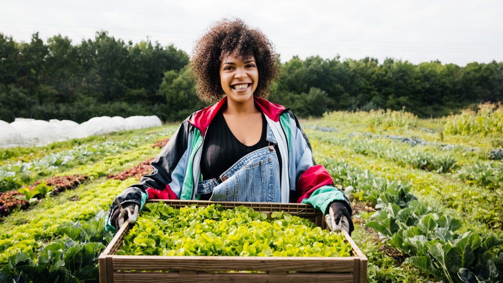 A smiling woman stands on a farm holding a crate of vegetables