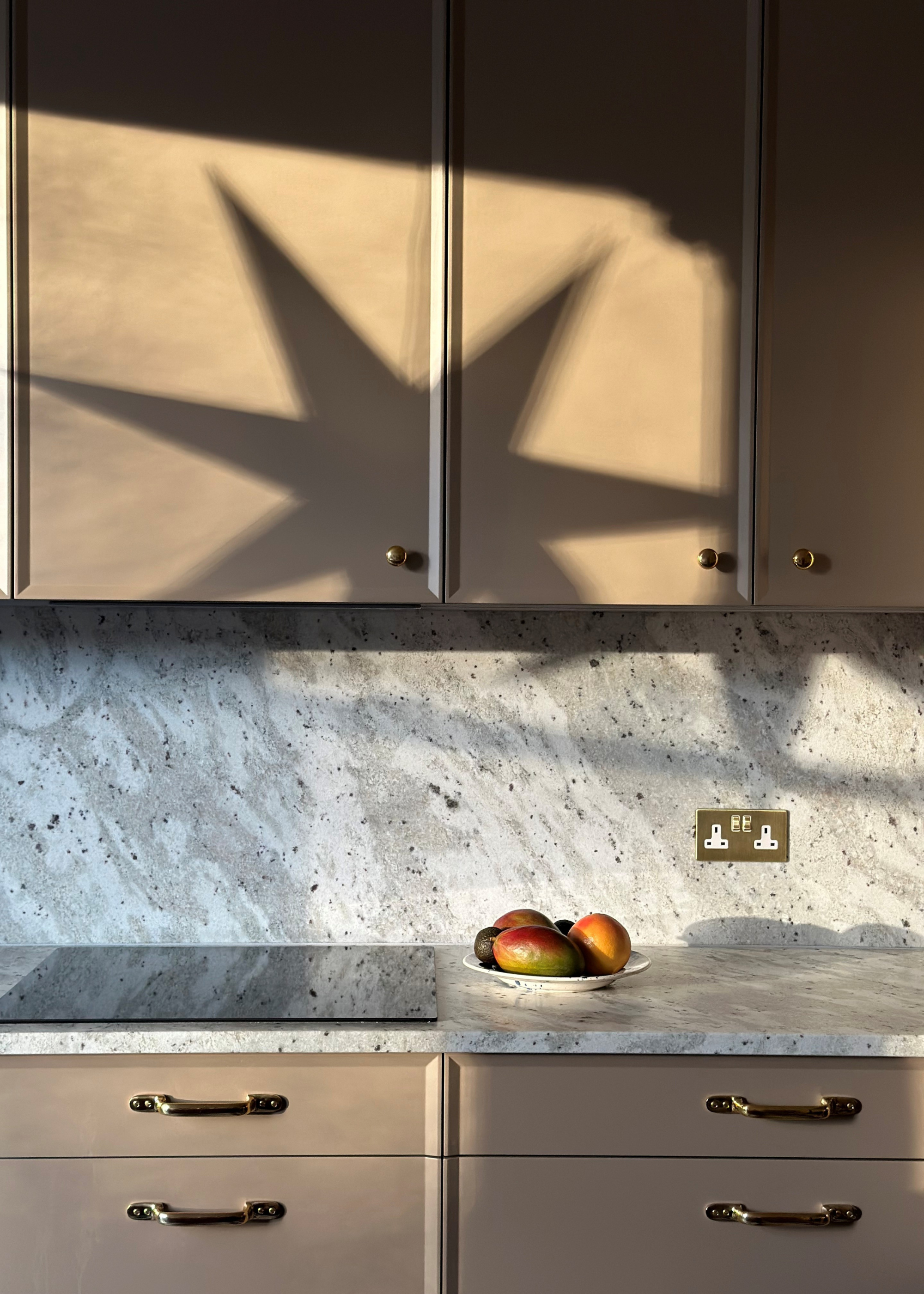 A kitchen with beige cabinets, a marble counter and backsplash, a bowl of mangoes, and the shadow of a star