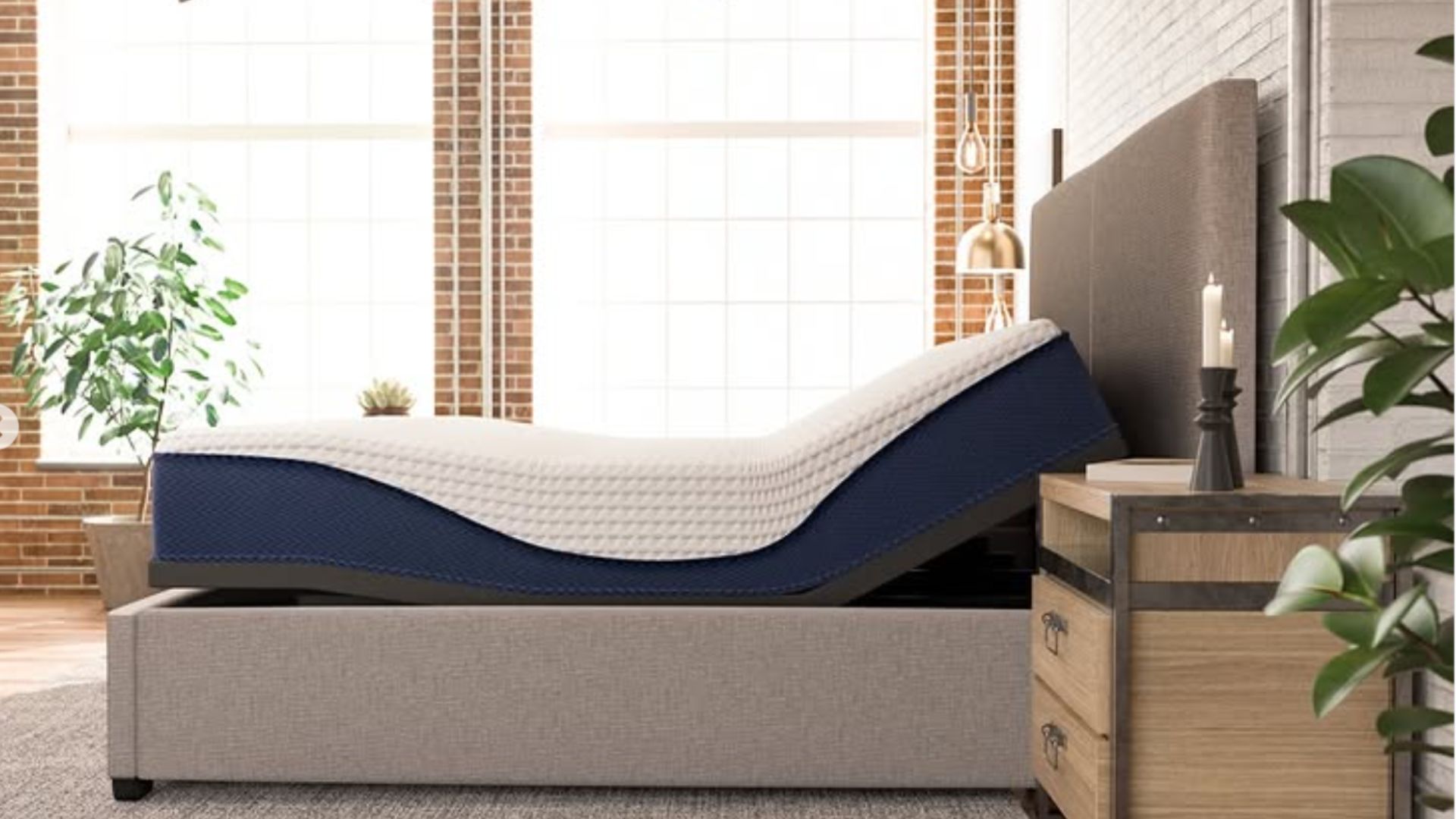 A dual-sided Sleep Number ComfortMode smart bed is shown in a modern, industrial-style bedroom with exposed brick walls and large windows. The bed features two independent mattresses on an adjustable base, with both the head and foot sections slightly elevated. The mattresses have a navy blue base with "ComfortMode" branding and a white, textured top cover. The bed is framed by a grey upholstered headboard and base, set on a grey area rug next to a wooden nightstand with minimalist decor.