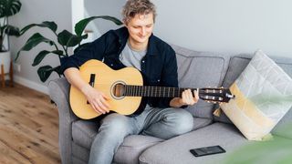 Man playing acoustic guitar with smartphone the sofa next to him