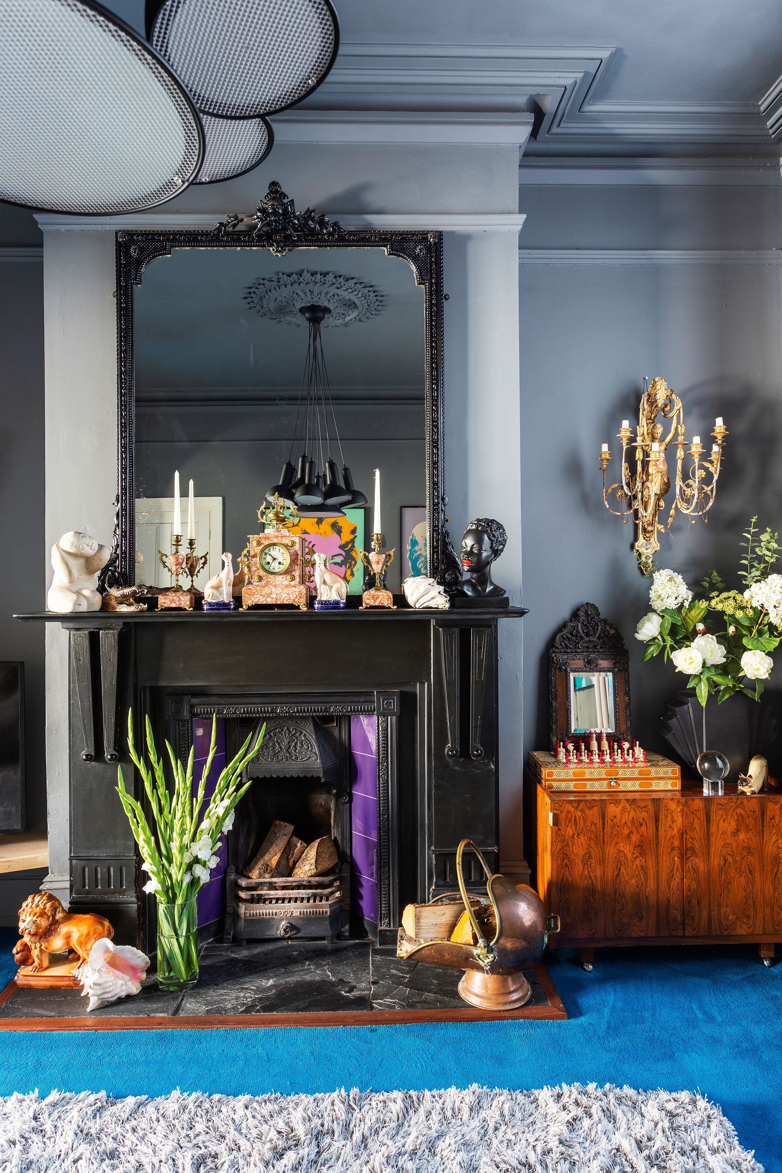 A grey living room with bright blue carpet, traditional black fireplace and large black framed mirror with chandelier ceiling light in reflection