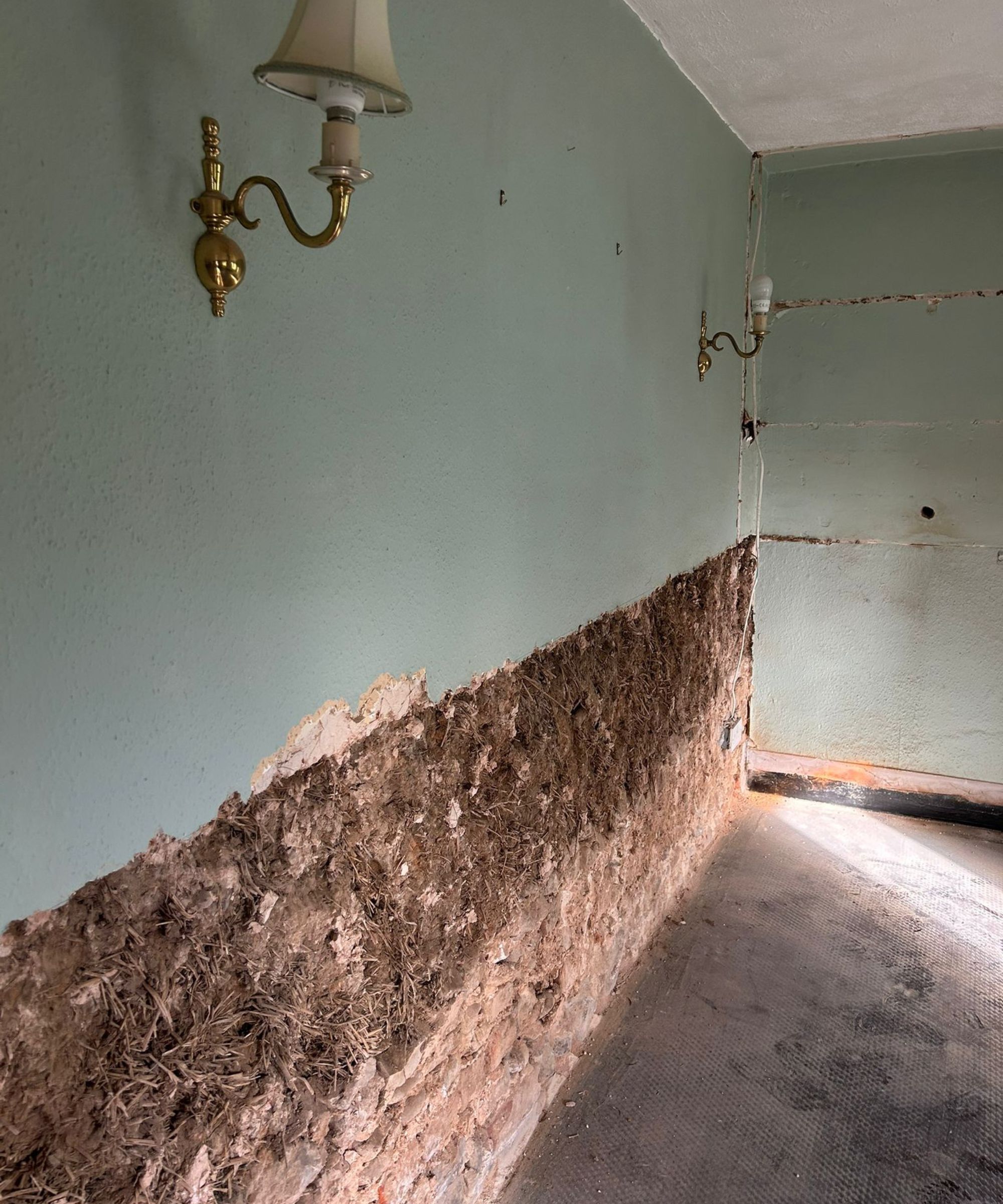 A partially stripped room with mint-green walls, where the lower plaster has been removed to expose rough cob and brickwork beneath. Gold wall sconces remain in place, and the bare floor adds to the raw, work-in-progress atmosphere.