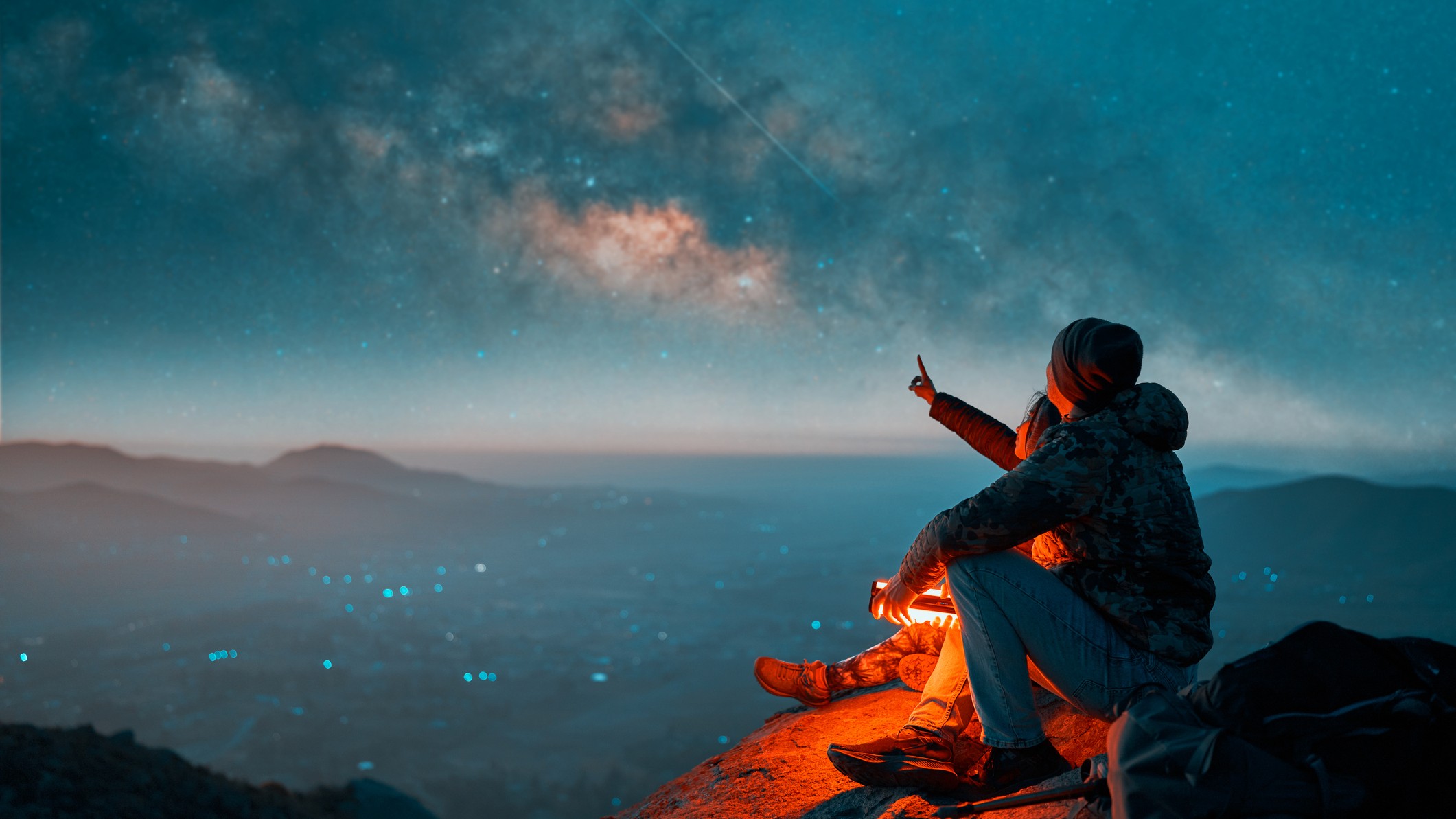 two people sit and look up at the night sky. one of them is pointing to the stars.