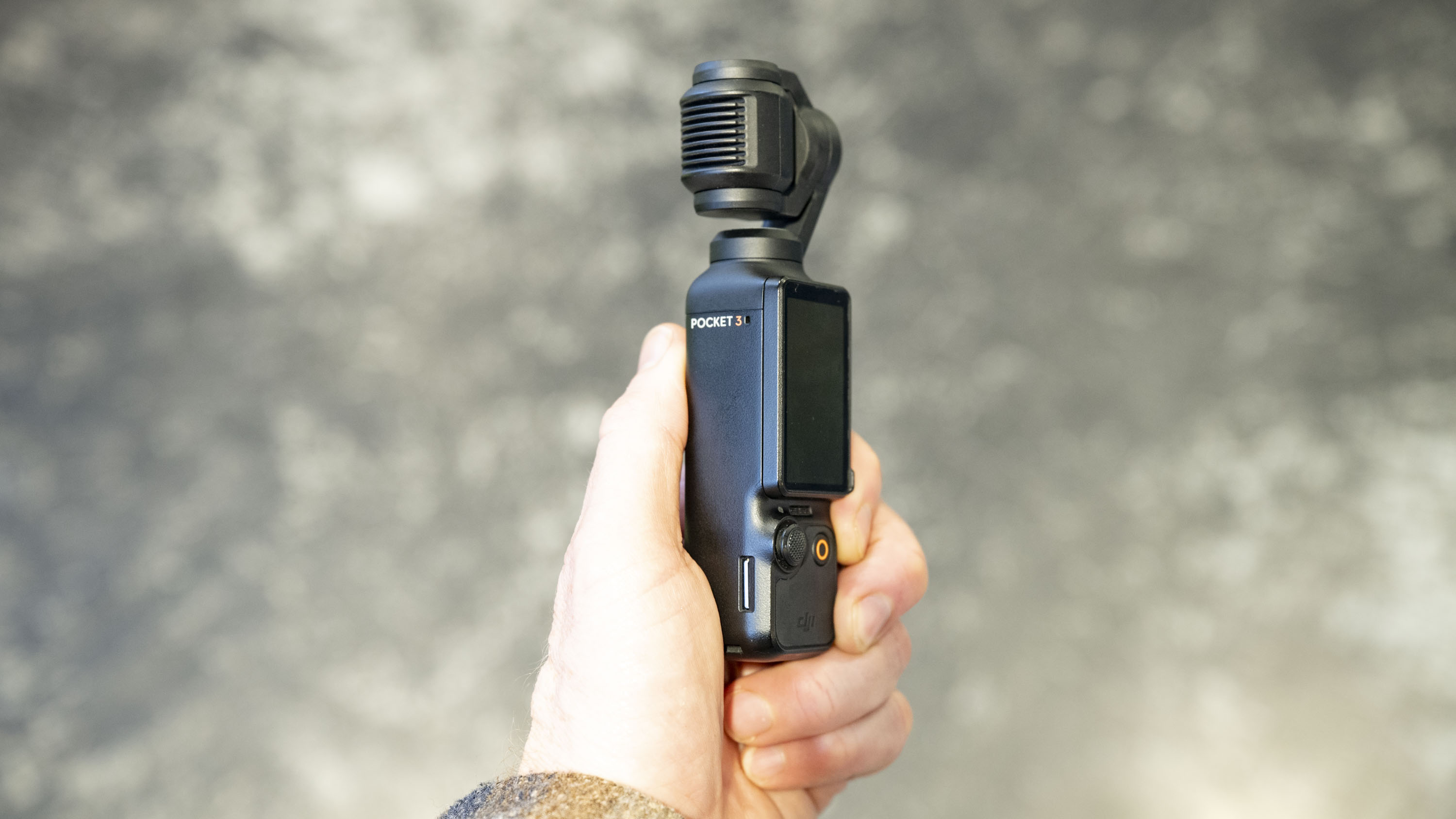 DJI Osmo Pocket 3 vlogging camera in user's hand, grey floor in background