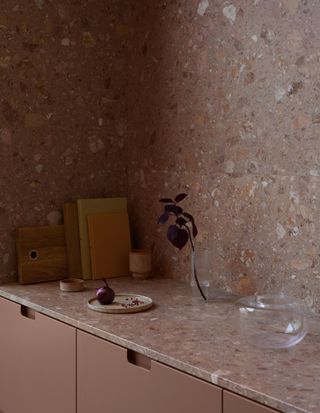 Image of a dusty pink cabinet and matching pink terrazzo countertop. The countertop extends into a high backsplash on two adjoining walls. In the back corner there are cutting boards and books stacked.