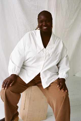 Model wears white button down shirt and brown pants while sitting on a stool like structure
