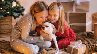 Two young girls surrounded by Christmas presents hugging their dog