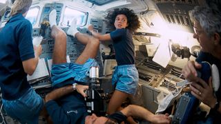 four astronauts in dark blue short sleeve polo shirts and light blue shorts work inside a space shuttle
