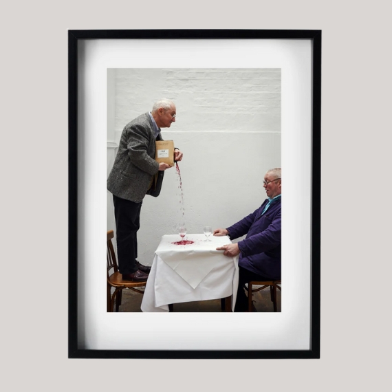 A photographic frame containing a photograph of two elderly man, one sitting on a chair with a blue jumper and glasses, the other standing on a chair while pouring wine, sits against a pale grey background.