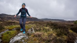 A woman stands on one foot on a mossy hillside, wearing a pair of Dynafit Feline SL Running Shoes.