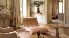A bright living room with aged mirrors, a pale pink lounger, a vase of stems, a chrome and pink table lamp, a wood side table, and long curtains