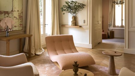 A bright living room with aged mirrors, a pale pink lounger, a vase of stems, a chrome and pink table lamp, a wood side table, and long curtains