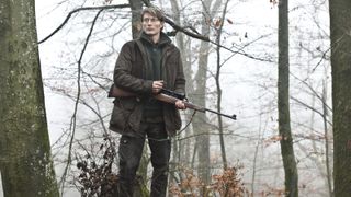Mads Mikkelsen står med ett gevär i handen ute i skogen i Jakten.