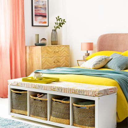 White bedroom with candy coloured accents, with an IKEA shelving unit used as a bench at the end of the bed 