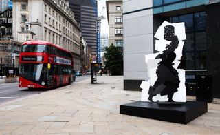 A fragmented monument depicting the silhouette of a female street vendor carring a burning brazier on her head. Placed on a black square platform on a pavement in front of a building and photographed during the day