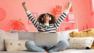 A young woman sitting on a sofa, wearing headphones and throwing her arms excitedly into the air.