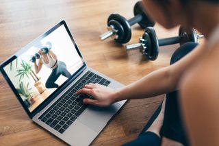 A woman watching a weight training video on her laptop with a pair of dumbbells.