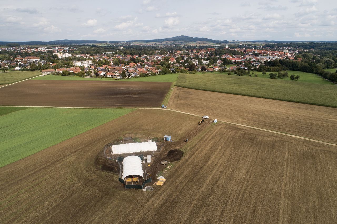 A bird's-eye view of the excavation site on a dirt and grass field with a village in the background.