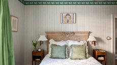 Bedroom with green wallpapered striped walls, an antique wooden headboard, white frilly bedding, green throw cushions, and two wooden nightstands with pleated sconces above.