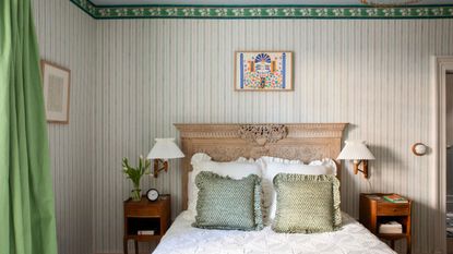 Bedroom with green wallpapered striped walls, an antique wooden headboard, white frilly bedding, green throw cushions, and two wooden nightstands with pleated sconces above.