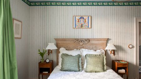 Bedroom with green wallpapered striped walls, an antique wooden headboard, white frilly bedding, green throw cushions, and two wooden nightstands with pleated sconces above.