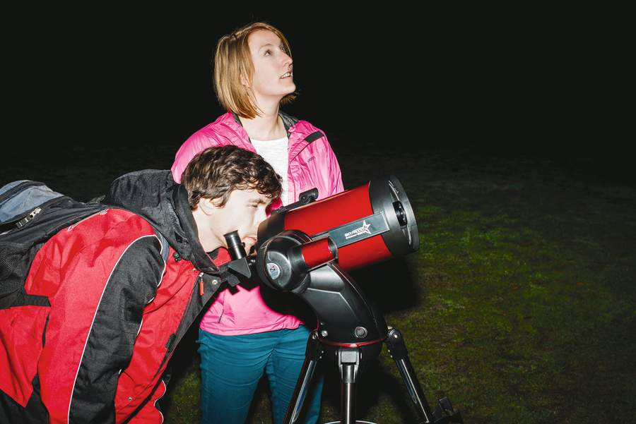 Two people stargazing. One is using a telescope and the other is just looking up at the sky. Shot outside in the dark.