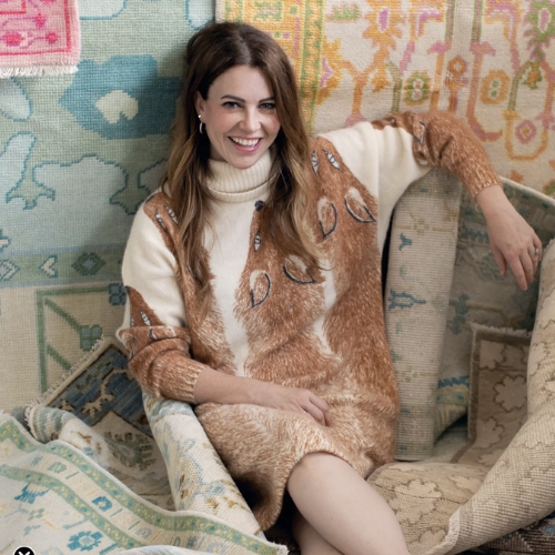 A woman in a turtleneck dress with foxes printed on it is sitting on a pile of colorful rugs.