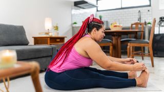 A woman performs a seated forward fold yoga move at home in her living room. She wears leggings and a vest top. She is on the floor, with her legs straight out and her torso leaning forward. She grasps her big toes with her hands.