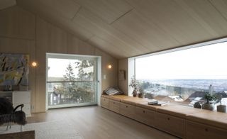 Alternative interior view of the main living area at the Oslo family house featuring wood panelled walls, wood flooring, a wood panelled ceiling, wall lights, wall art, an off-white coloured rug, seating and large windows offering views of Oslo