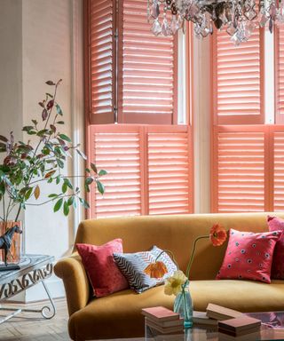Coral colored window shutters in living room with mustard velvet upholstered sofa and chandelier light fixture