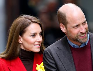 Kate Middleton wearing a red coat with a yellow daffodil pin looking to the right and smiling standing next to Prince William, looking in the same direction