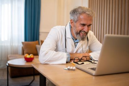 An older doctor smiles into a laptop during a telehealth video chat with a patient.