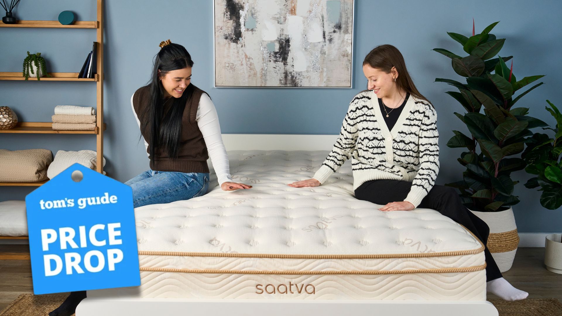 Two girls sitting on white and gold Saatva Classic mattress in sleep studio with blue wall, canvas, plants and shelving next to bed 